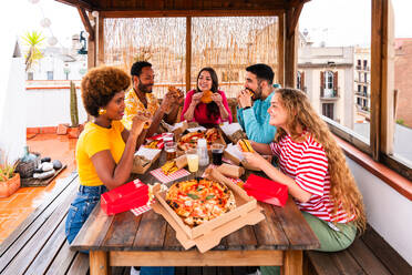 Multiethnic group of happy young friends having dinner barbecue party on rooftop at home - Multiracial cheerful young adult people having fun and bonding on a terrace balcony with city view, eating and drinking. - DMDF09184