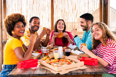 Multiethnic group of happy young friends having dinner barbecue party on rooftop at home - Multiracial cheerful young adult people having fun and bonding on a terrace balcony with city view, eating and drinking. - DMDF09185