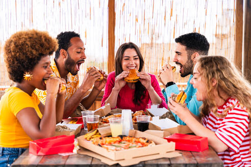 Multiethnic group of happy young friends having dinner barbecue party on rooftop at home - Multiracial cheerful young adult people having fun and bonding on a terrace balcony with city view, eating and drinking. - DMDF09189