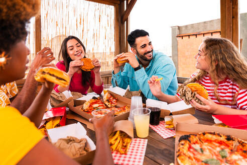 Multiethnic group of happy young friends having dinner barbecue party on rooftop at home - Multiracial cheerful young adult people having fun and bonding on a terrace balcony with city view, eating and drinking. - DMDF09194