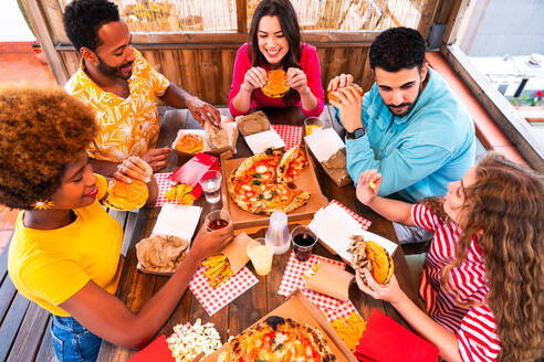 Multiethnic group of happy young friends having dinner barbecue party on rooftop at home - Multiracial cheerful young adult people having fun and bonding on a terrace balcony with city view, eating and drinking. - DMDF09195