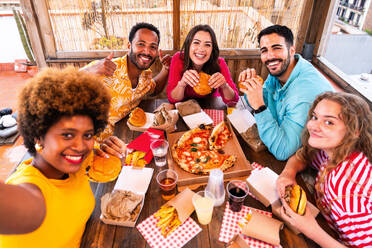 Multiethnic group of happy young friends having dinner barbecue party on rooftop at home - Multiracial cheerful young adult people having fun and bonding on a terrace balcony with city view, eating and drinking. - DMDF09196