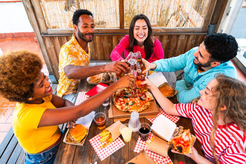 Multiethnic group of happy young friends having dinner barbecue party on rooftop at home - Multiracial cheerful young adult people having fun and bonding on a terrace balcony with city view, eating and drinking. - DMDF09197