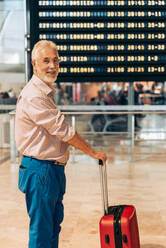 Side view of anonymous senior male traveler with gray hair looking at camera board and standing with suitcase at modern aerodrome while going on vacation - ADSF51154