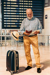 Full body of bearded smiling senior male passenger checking passport and boarding pass while standing with suitcase and hat against timetable board at airport - ADSF51155