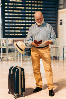 Full body of bearded smiling senior male passenger checking passport and boarding pass while standing with suitcase and hat against timetable board at airport - ADSF51155