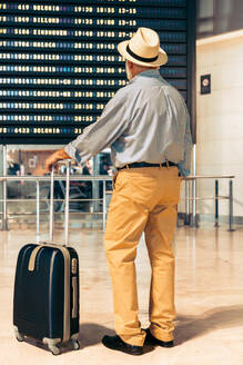 Back view of anonymous senior male traveler with gray hair looking at departure board and standing with suitcase at modern aerodrome while going on vacation - ADSF51156