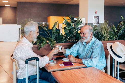 Happy senior male friends dressed in casual clothes toasting coffee cups and sitting at wooden table while waiting for flight at airport cafeteria - ADSF51158