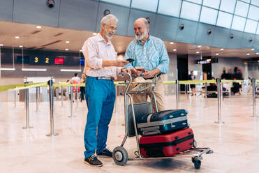 Happy senior male passenger friends in casual clothes checking boarding pass in mobile while standing against timetable and waiting for departure at modern airport - ADSF51161