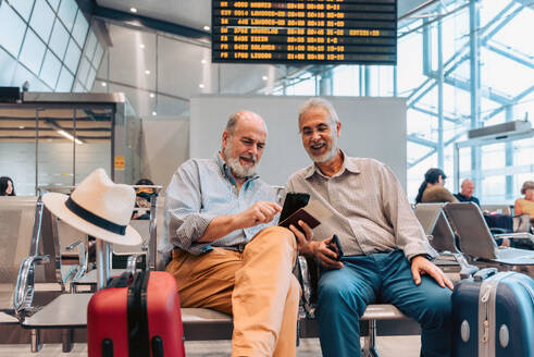 Happy senior male tourists dressed in casual clothes sitting with suitcase on chairs and using mobile phones while waiting in contemporary airport lounge - ADSF51162