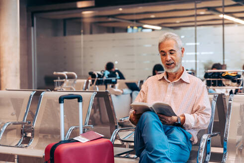Bearded focused senior male passenger in eyeglass and casual attire sitting with suitcase of chair and reading book while waiting for flight at airport lounge - ADSF51164