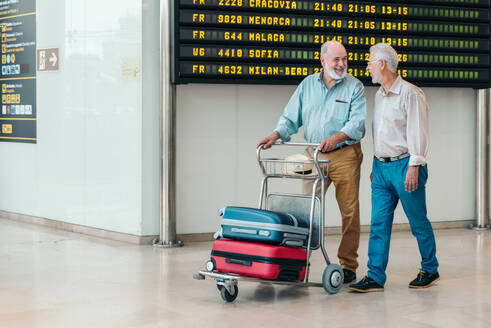 Happy senior male tourists in casual clothes holding suitcase and walking from shore to against flight timetable board at modern airport - ADSF51165