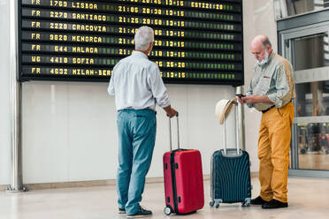 Back view of anonymous senior male tourist in casual attire looking at departure schedule board at contemporary airport while his friend texts on his mobile phone while going on vacation - ADSF51166