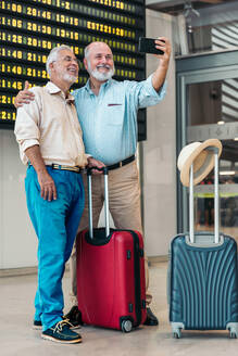 Full body of positive senior male friends dressed in casuals taking selfie over smartphone while standing with suitcases against departure board at airport - ADSF51169