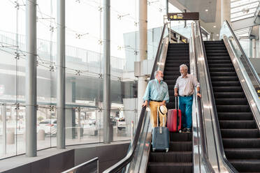 Low angle view of senior male travelers in casuals talking with each other while standing with luggage on moving stairs at airport terminal - ADSF51171