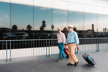 Full body of senior friends dressed in casuals holding suitcase and walking on footpath by railing while seek for taxi outside Valencia airport - ADSF51174