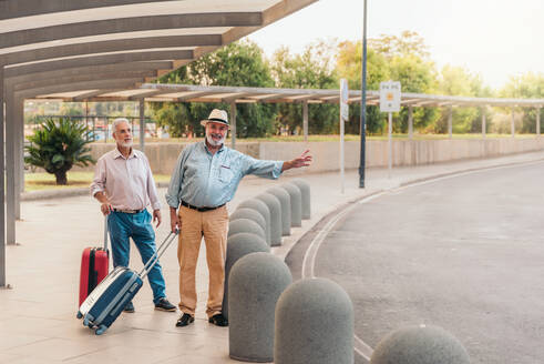 Full body of smiling senior man with hat and suitcase hailing for cab while standing with friend on footpath outside airport terminal during vacation - ADSF51176