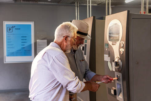 Focused senior male passengers dressed in casual clothes inserting credit card for withdrawing cash from ATM machine at airport terminal - ADSF51178