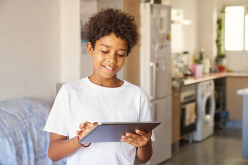 Smiling African American teenager boy with curly dark hair and in casuals using digital tablet while standing in living room at home - ADSF51183