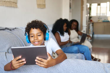 Smiling teenager wearing headphones and watching movie on digital tablet while sitting on sofa with mother and grandmother in background - ADSF51189