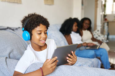 Smiling teenager wearing headphones and watching movie on digital tablet while sitting on sofa with mother and grandmother in background - ADSF51190