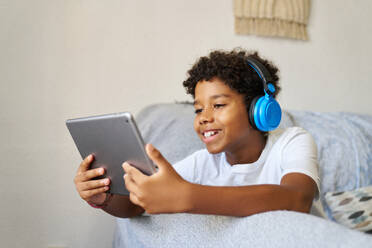 Happy African American teen boy with curly black hair wearing headphones and using digital tablet while relaxing on sofa in living room - ADSF51191