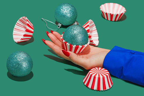 A hand with red nails holding a glittering turquoise bauble in a striped cupcake liner, with a festive feel. - ADSF51197
