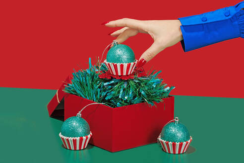 A hand in a blue sleeve reaches for a glittery green Christmas ornament cupcake atop festive tinsel in a red box - ADSF51198