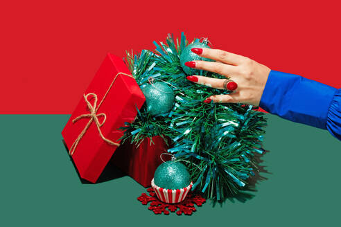 A hand from a blue sleeve delicately touches Christmas baubles nestled in tinsel inside a red gift box - ADSF51200