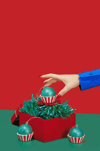 A hand in a blue jacket positions a green glitter Christmas cupcake ornament into a red box with tinsel - ADSF51201