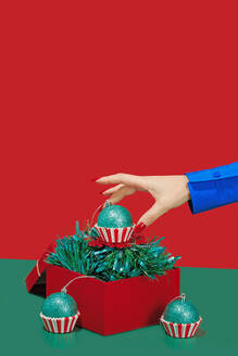 A hand in a blue jacket positions a green glitter Christmas cupcake ornament into a red box with tinsel - ADSF51201