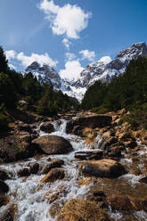 Pristine mountain stream flowing through rocky terrain with snow-capped peaks in the background, under a blue sky with feathery clouds - ADSF51231