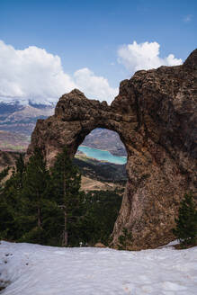 A natural rock arch with a stunning view of a turquoise mountain lake, set against the snow-capped Pyrenees in Ordesa National Park - ADSF51235