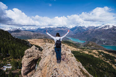 A woman stands triumphant on a rocky ridge with outstretched arms, overlooking a scenic vista of mountains and a lake in Ordesa National Park, Pyrenees - ADSF51236