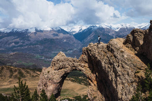 Adventurous hiker standing on a natural stone arch with breathtaking views of the snow-capped Pyrenees mountains near Torla, in Ordesa National Park - ADSF51237