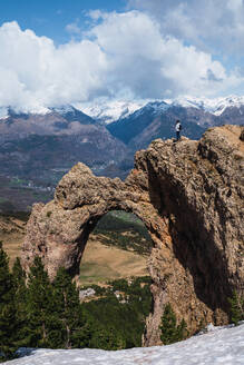 A solitary hiker stands atop a rugged formation, admiring the expansive view of snow-covered peaks through a unique natural stone arch in the Pyrenees - ADSF51238