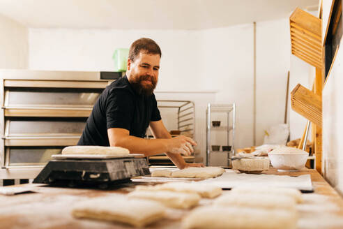 Focused bearded Caucasian male baker dressed in casuals kneading raw dough on table for making pastry in kitchen at bakehouse - ADSF51239