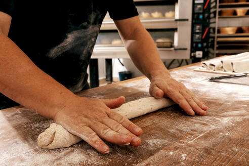 Crop hands of anonymous male cook in casuals kneading dough on wooden table with flour while preparing bread in kitchen of bakehouse - ADSF51243