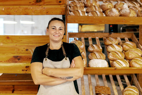 Portrait of confident positive female seller in apron with arms crossed standing by freshly baked breads arranged on wooden shelves in bakery - ADSF51252