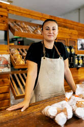 Portrait of confident positive female seller in apron with his hands on the counter standing by freshly baked breads arranged on wooden shelves in bakery - ADSF51254