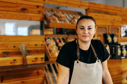 Portrait of confident positive female seller in apron with his hands on the counter standing by freshly baked breads arranged on wooden shelves in bakery - ADSF51255