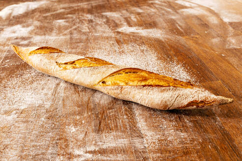 Closeup of freshly baked delicious baguette with crunchy crust placed on messy wooden table in kitchen at bakehouse - ADSF51256