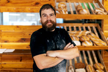 Portrait of confident positive bearded mature man with arms crossed standing against wooden shelves with freshly baked breads in bakery - ADSF51257