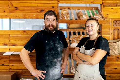 Positive male and female coworkers in apron and casuals looking at camera while standing against wooden shelves with baked items in bakery - ADSF51258