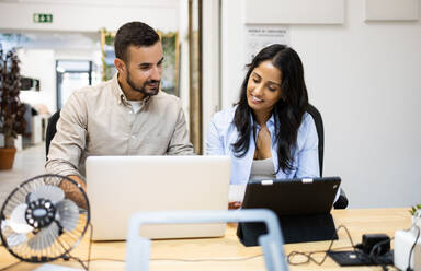 Smiling multiracial male and female colleagues discussing plan over digital tablet and laptop while sitting at desk in creative office - ADSF51262