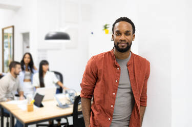 Portrait of confident African American young businessman wearing casuals standing against wall and looking at camera in creative office - ADSF51267