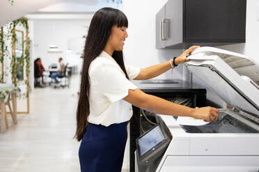 Side view of smiling Asian young businesswoman in casuals with long hair using modern copier while working in creative office - ADSF51269