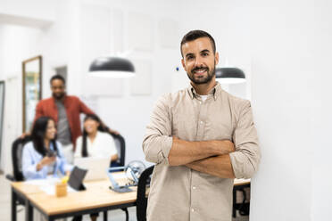 Portrait of smiling confident young Caucasian businessman in casuals with arms crossed leaning on wall in creative office - ADSF51270