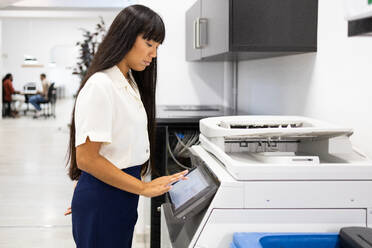 Side view of serious Asian young businesswoman in casuals with long hair using modern copier while working in creative office - ADSF51272