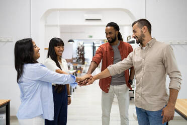 Cheerful multiracial male and female colleagues dressed in casuals stacking hands together while standing in creative office - ADSF51273